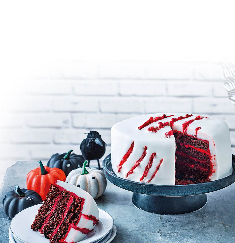 Das Bild zeigt eine mehrschichtige Torte auf einer schwarzen Tortenplatte. Die Torte ist au en mit glattem, wei em Fondant berzogen. Auf der Oberfl che und an der Seite sind rote Linien und Risse zu sehen, die an Kratzspuren erinnern und der Torte ein gruseliges Halloween-Design verleihen. Im Anschnitt erkennt man mehrere dunkle Schokoladenb den, die mit einer leuchtend roten Creme geschichtet sind. Daneben steht ein Teller mit einem St ck der Torte. Im Hintergrund sind kleine Zierk rbisse in Wei , Schwarz und Orange zu sehen – passend zur herbstlichen Halloween-Stimmung.