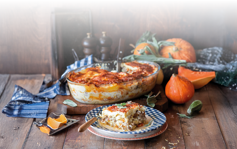 Das Bild zeigt eine herbstlich angerichtete K rbis-Lasagne – k stlich berbacken mit goldbrauner K sekruste. Im Vordergrund liegt ein angerichtetes St ck auf einem Teller mit blau-wei em Muster, daneben ein rustikales Holzmesser. Im Hintergrund stehen K rbisse, frische Salbeibl tter und eine Auflaufform, aus der bereits Portionen entnommen wurden.