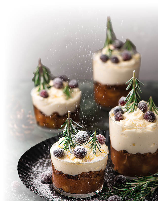 Vier Gl ser mit Schichtdessert aus brauner Creme und wei er Sahne, garniert mit Rosmarinzweigen und roten Beeren, dar ber f llt Puderzucker.