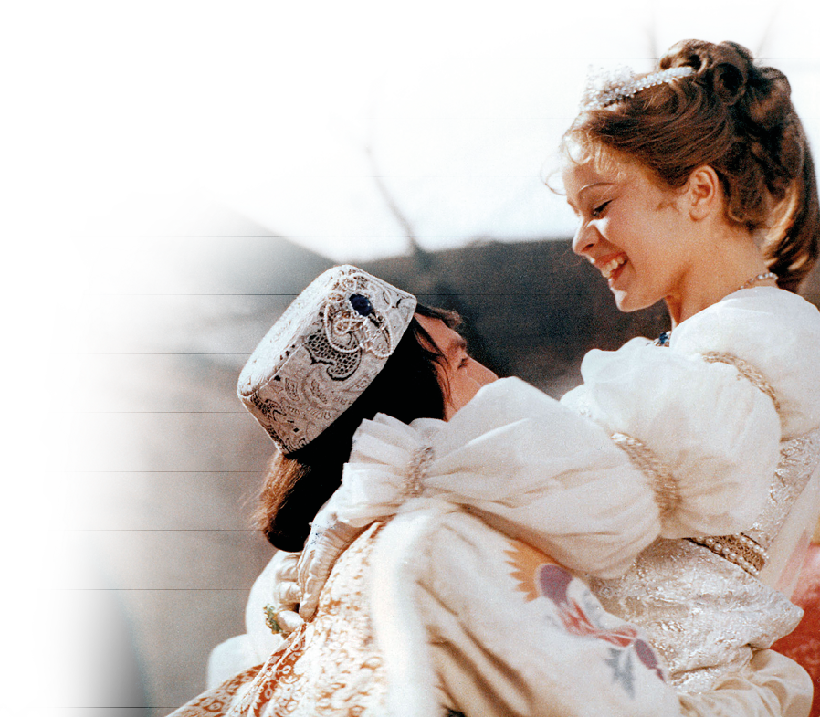 Auf dem Bild sieht man eine winterliche, m rchenhafte Szene: Eine junge Frau und ein junger Mann – beide warm in festliche, helle Winterkleidung geh llt – stehen sich freudestrahlend gegen ber. Der Mann hebt die junge Frau leicht, beide lachen und wirken vertraut und gl cklich. Der Hintergrund ist weich verschwommen, was den m rchenhaften Eindruck noch verst rkt.   Es handelt sich um den Weihnachtsfilmklassiker „Drei Haseln sse f r Aschenbr del“.