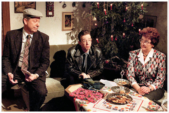 Auf dem Bild sitzen drei Personen in einem Wohnzimmer, das weihnachtlich dekoriert ist.   Links sitzt ein Mann mit Schirmm tze und Mantel. Er wirkt eher zur ckhaltend oder nachdenklich.    In der Mitte sitzt ein j ngerer Mann in einer dunklen Lederjacke, der leicht nach vorn gebeugt ist und aufmerksam in die Runde schaut.    Rechts sitzt eine Frau mit rot gelocktem Haar und gemusterter Jacke. Sie schaut zur Seite und wirkt, als w re sie in ein Gespr ch vertieft.    Im Hintergrund steht ein geschm ckter Weihnachtsbaum mit Kerzen und Lametta. Auf dem Tisch vor ihnen liegen Geb ck, eine brennende Kerze sowie verschiedene Teller – es sieht nach einem klassischen, etwas nostalgischen Weihnachtsszenario aus. Es ist eine Szene aus dem Weihnachtssketch “Familie Heinz Becker".