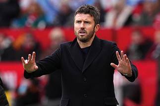 Mann in schwarzem Mantel mit erhobenen H nden vor unscharfem Hintergrund mit Menschen und rotem Banner