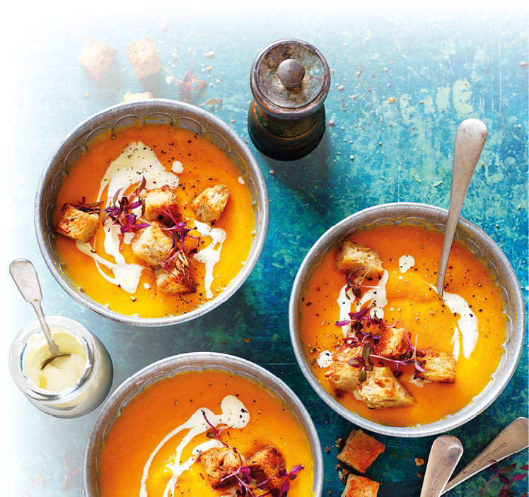 Drei Schalen mit cremiger Suppe, garniert mit Croutons, Sahne und Kr utern, auf blauem Untergrund mit zwei L ffeln und einem Pfefferstreuer.