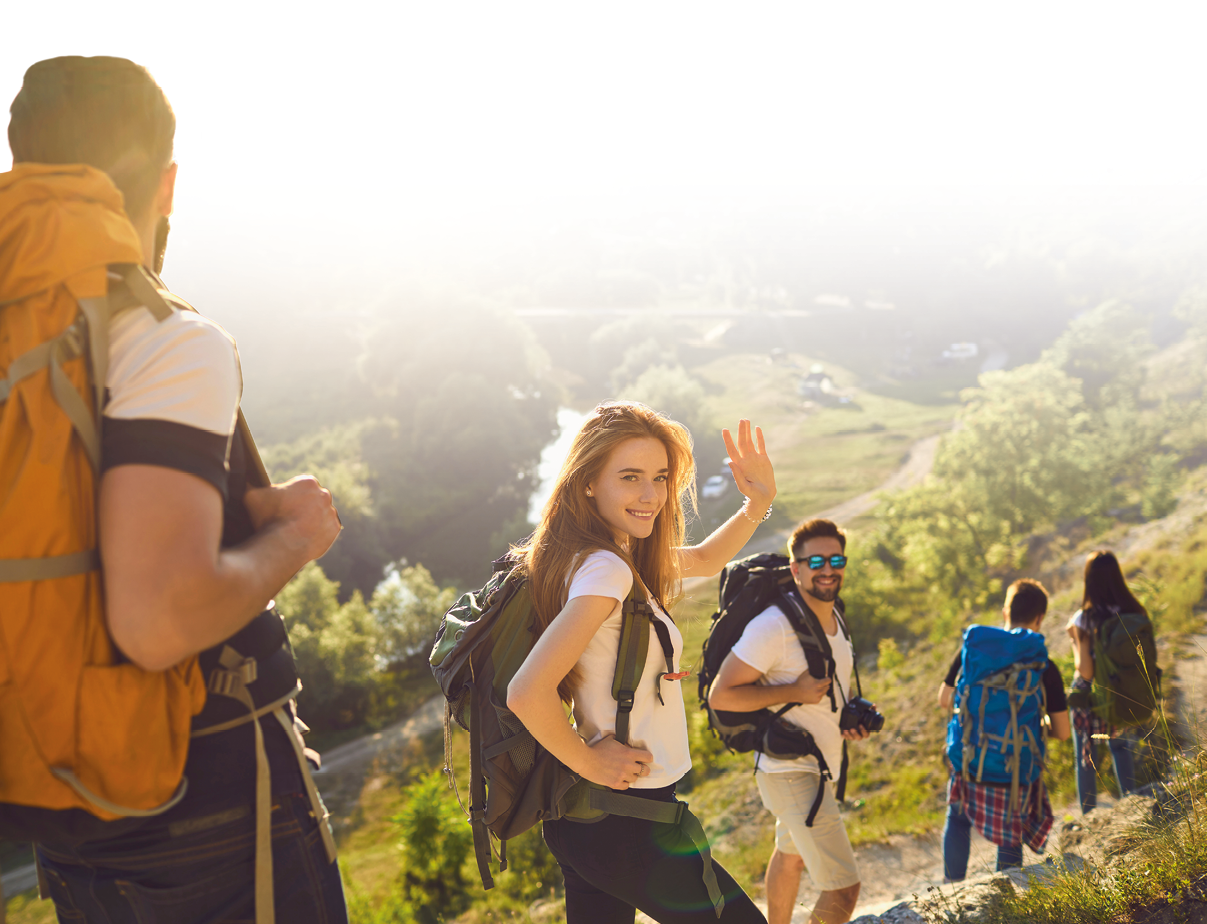 Group of happy backpackers trekking on sunny day. Young tourists traveling and enjoying active summer vacation. Smiling woman looking at camera and waving hand walking down hiking trail with friends