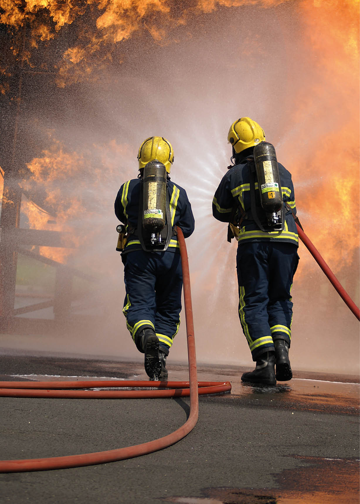 Zwei Personen in Feuerwehrschutzkleidung l schen mit Schl uchen ein gro es Feuer vor einem brennenden Geb ude.   Beschreibung von KI generiert