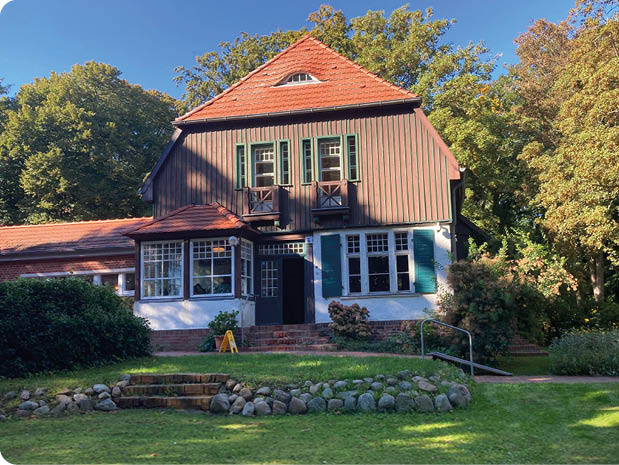 Ein zweist ckiges Haus mit rotem Ziegeldach steht in einem gr nen Garten mit B umen und einem gepflegten Rasen unter klarem blauem Himmel. Beschreibung von KI generiert