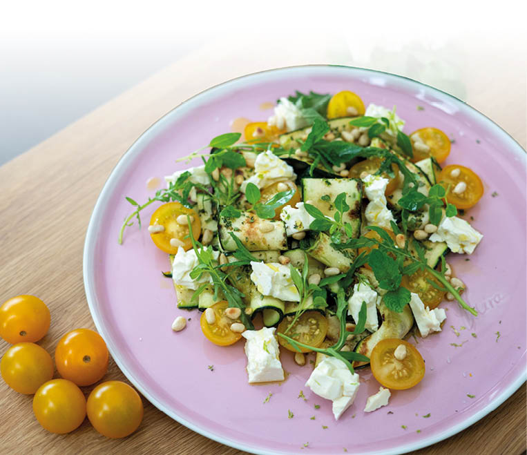 Auf einem rosa Teller liegt ein Salat aus gelben Kirschtomaten, wei en K sew rfeln, gr nen Kr utern und Pinienkernen, daneben liegen mehrere gelbe Kirschtomaten auf einem Holzbrett. Beschreibung von KI generiert