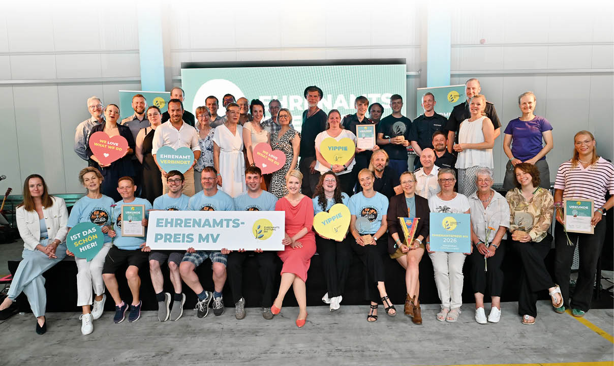 Eine gro e Gruppe von Erwachsenen und Senioren sitzt und steht in einem hellen Raum zusammen und h lt bunte Schilder mit positiven Botschaften und einem Banner mit der Aufschrift Ehrenamtspreis MV. Beschreibung von KI generiert