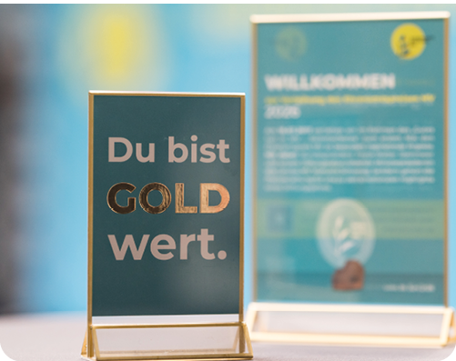 Ein Schild mit dem Text Du bist GOLD wert steht vor einem unscharfen Hintergrund mit einem weiteren Schild und einem hellblauen Farbverlauf. Beschreibung von KI generiert