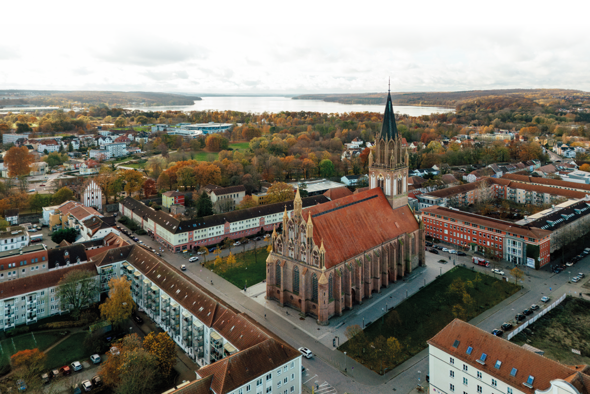 Eine gro e gotische Kirche mit rotem Dach steht inmitten eines Wohngebiets mit herbstlich gef rbten B umen und einem Fluss im Hintergrund. Beschreibung von KI generiert