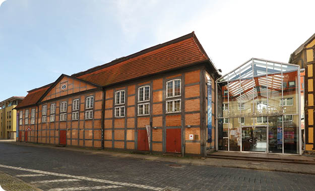 Ein historisches Fachwerkgeb ude mit roten T ren und Fenstern steht neben einem modernen Glasbau in einer ruhigen Stra enumgebung. Beschreibung von KI generiert