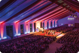 In einer gro en Halle mit farbiger Beleuchtung spielt ein Orchester vor einem Publikum, das auf Stuhlreihen sitzt. Beschreibung von KI generiert