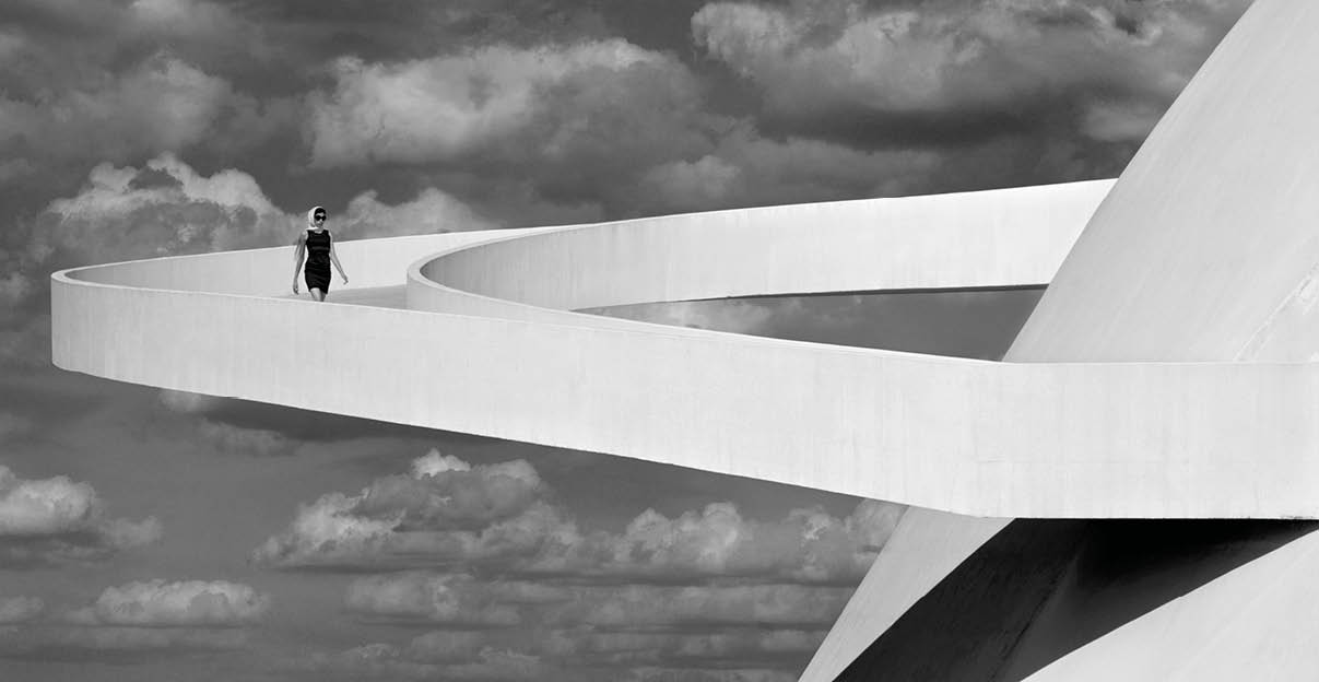 Eine Person in dunkler Kleidung geht auf einem breiten, geschwungenen, wei en Weg, der sich vor einem dramatischen Wolkenhimmel und einer modernen architektonischen Struktur erstreckt. Beschreibung von KI generiert