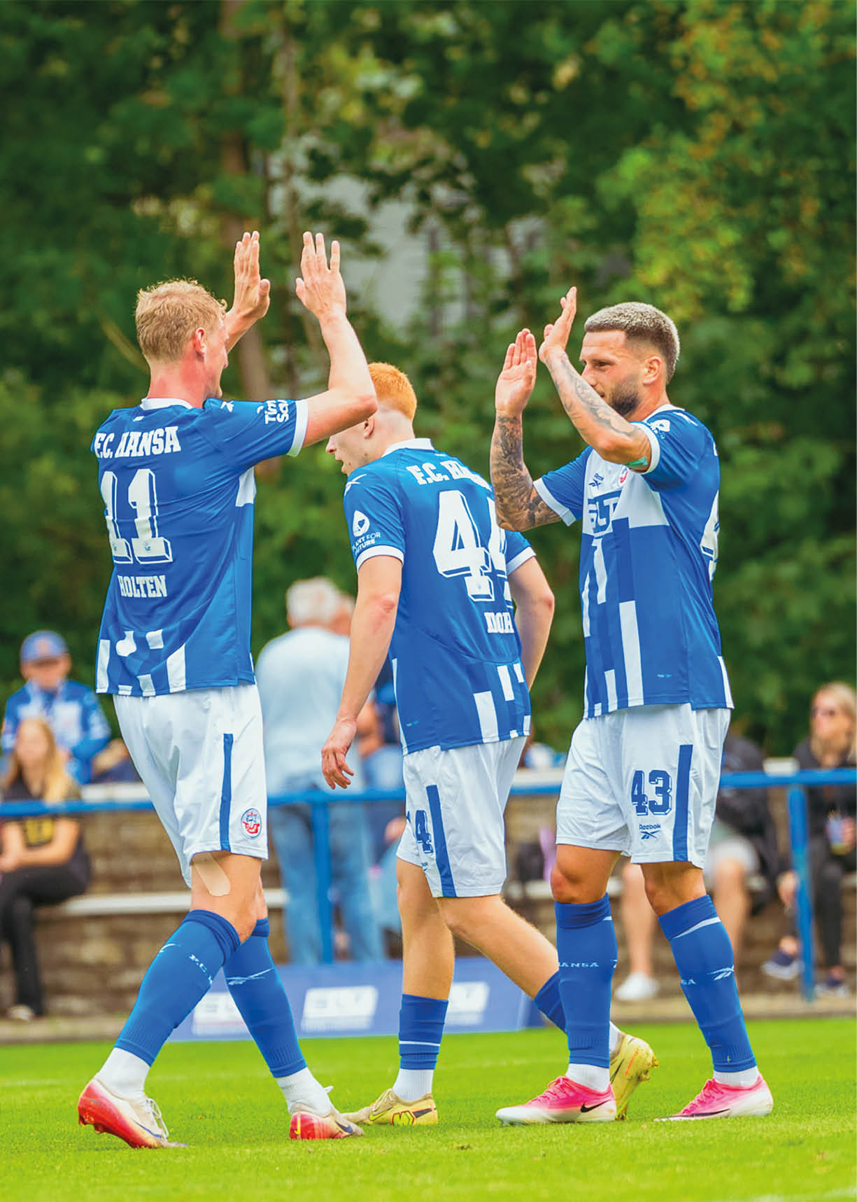 Drei Personen in blauen Fu balltrikots feiern auf einem Spielfeld mit erhobenen H nden, w hrend im Hintergrund Zuschauer auf einer Trib ne sitzen. Beschreibung von KI generiert