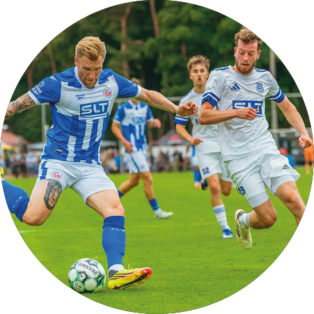 Eine Person in blau-wei em Sporttrikot und Shorts spielt aktiv Fu ball auf einem Spielfeld mit weiteren Personen im Hintergrund. Beschreibung von KI generiert