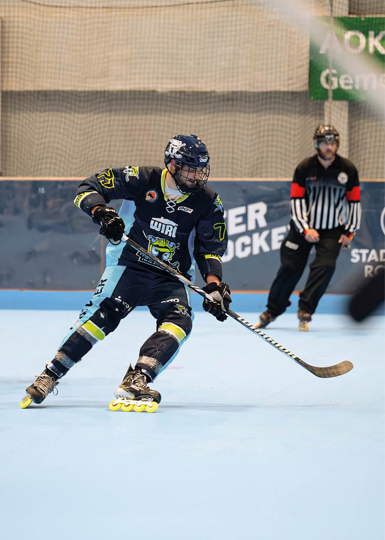 Eine Person in Schutzkleidung und Helm f hrt auf Inline-Skates und h lt einen Hockeyschl ger auf einem Hallenboden, w hrend eine weitere Person im Hintergrund eine Schiedsrichteruniform tr gt. Beschreibung von KI generiert