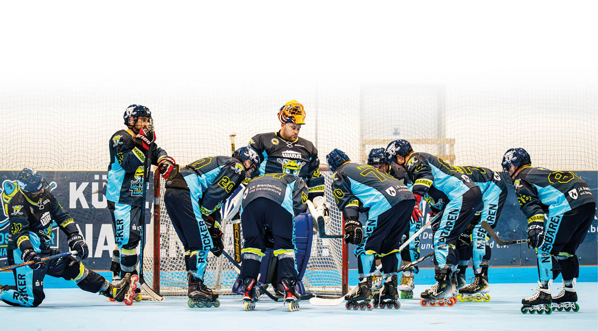 Eine Gruppe von Personen in Sportkleidung und Inline-Skates steht in einer Halle auf einem Spielfeld und bereitet sich auf ein Spiel vor, wobei sie sich in einer Reihe vor einem Tor versammeln. Beschreibung von KI generiert