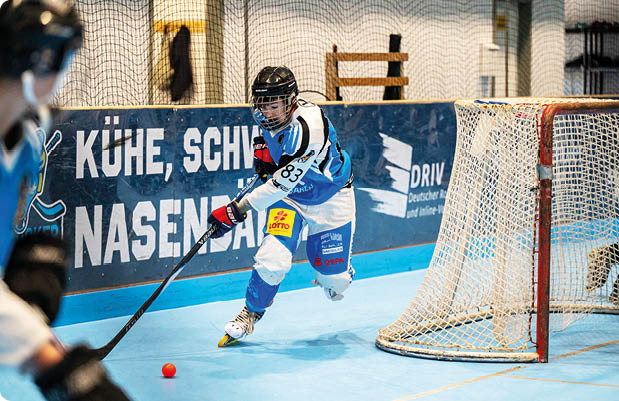 Person in Sportkleidung und Helm spielt mit Schl ger und Ball in einer Halle vor einem Tor beim Hallenhockey oder Inlinehockey. Beschreibung von KI generiert