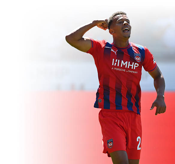 Eine Person in einem rot-blauen Sporttrikot und roten Shorts feiert auf einem Spielfeld mit erhobenem Arm und zeigt eine energische Geste. Beschreibung von KI generiert