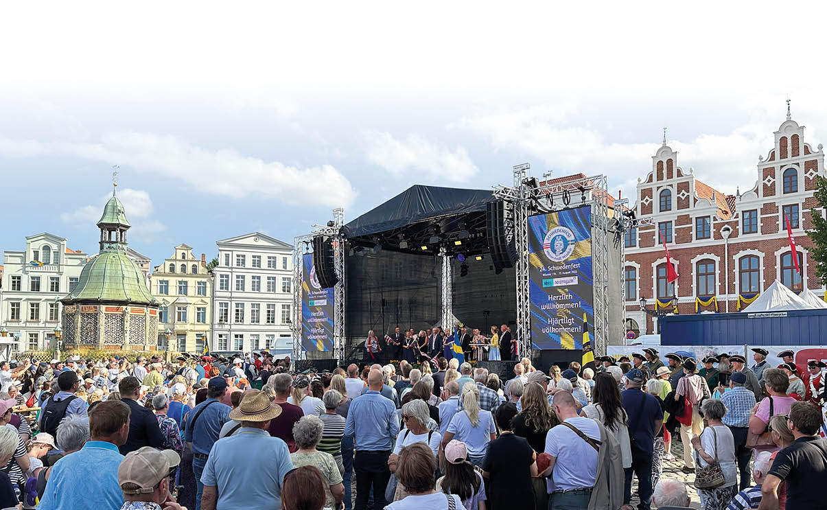 Eine gro e Menschenmenge steht auf einem Platz vor einer B hne mit Musikern, umgeben von historischen Geb uden bei Tageslicht. Beschreibung von KI generiert