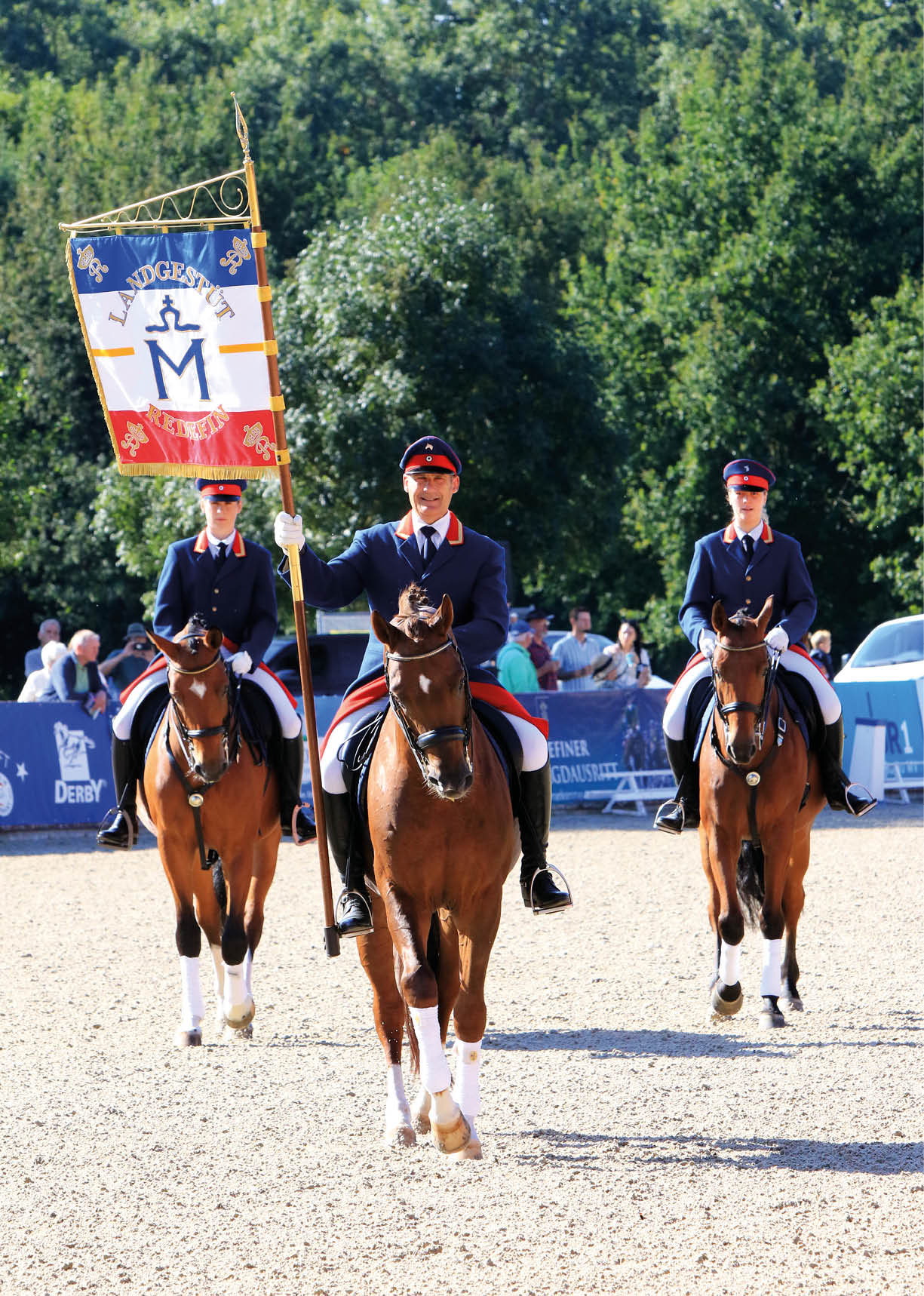 Drei Personen in formeller Reitkleidung reiten auf Pferden in einer sonnigen Arena, wobei die mittlere Person eine Fahne mit einem Wappen h lt. Beschreibung von KI generiert