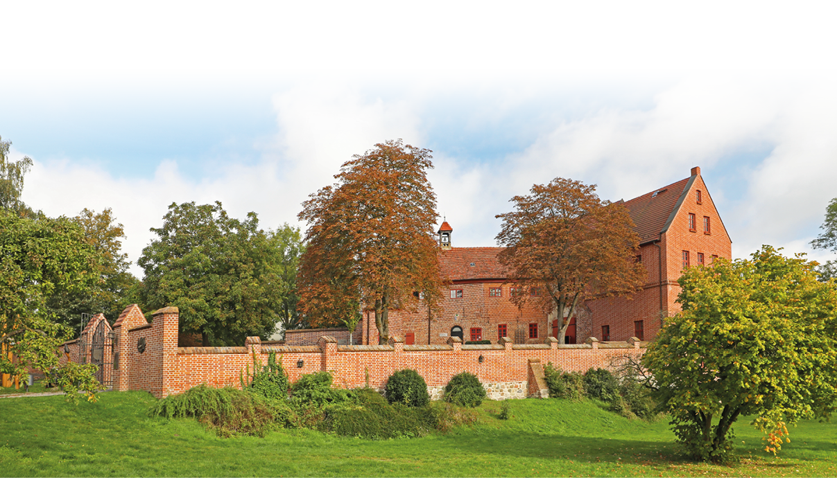 Ein historisches Backsteingeb ude mit Zinnenmauer steht in einer gr nen Parklandschaft unter einem blauen Himmel mit vereinzelten Wolken. Beschreibung von KI generiert