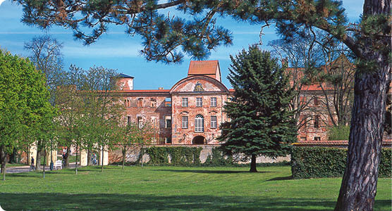Ein gro es historisches Geb ude aus rotem Backstein steht hinter einem weitl ufigen gr nen Rasen mit B umen unter einem blauen Himmel. Beschreibung von KI generiert