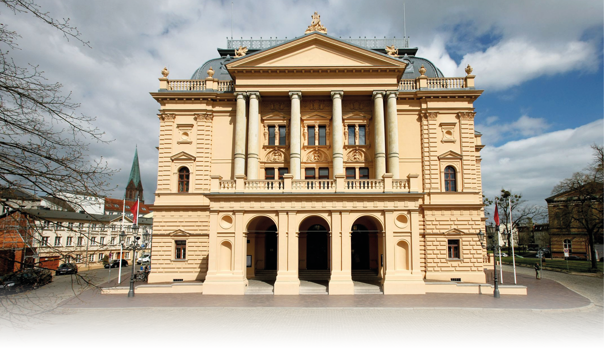 Ein historisches, klassizistisches Geb ude mit S ulen und drei gro en Torb gen steht unter einem bew lkten Himmel in einer st dtischen Umgebung. Beschreibung von KI generiert