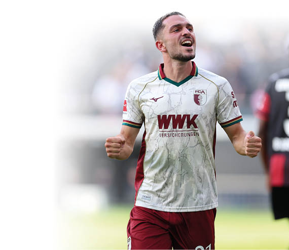 Ein jubelnder Spieler in wei -rotem Fu balltrikot steht auf dem Spielfeld, w hrend eine weitere Person in schwarzem Trikot mit der Nummer drei im Hintergrund zu sehen ist. Beschreibung von KI generiert