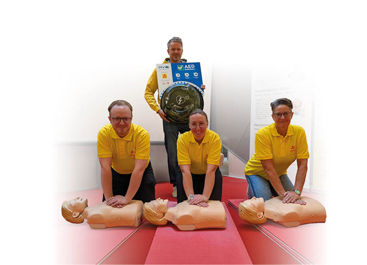 Vier Personen in gelben Poloshirts ben an drei  bungspuppen Herz-Lungen-Wiederbelebung, w hrend eine weitere Person im Hintergrund ein Defibrillator-Set h lt.  Beschreibung von KI generiert