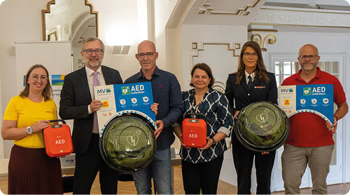 Sechs Erwachsene stehen in einem hellen Raum und halten Defibrillatoren und Informationsplakate zu einem Erste-Hilfe-Thema. Beschreibung von KI generiert