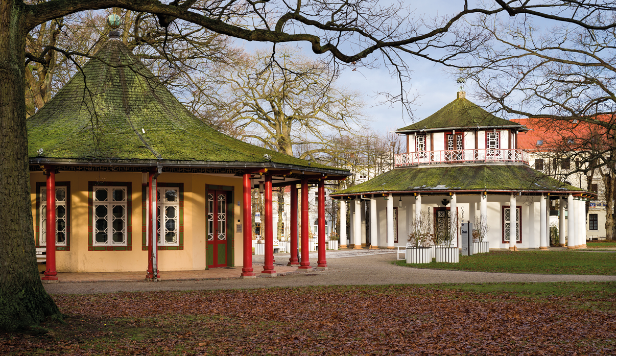 Zwei historische Pavillons mit spitzen D chern und roten S ulen stehen in einem Park mit kahlen B umen und herbstlichem Laub auf dem Boden. Beschreibung von KI generiert