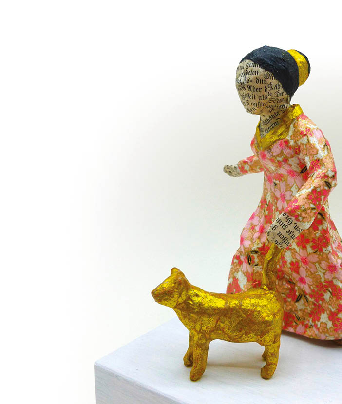 Eine bemalte Papierfigur in einem lang rmligen Kleid mit Blumenmuster h lt an einer Leine eine goldene Katze. Beschreibung von KI generiert